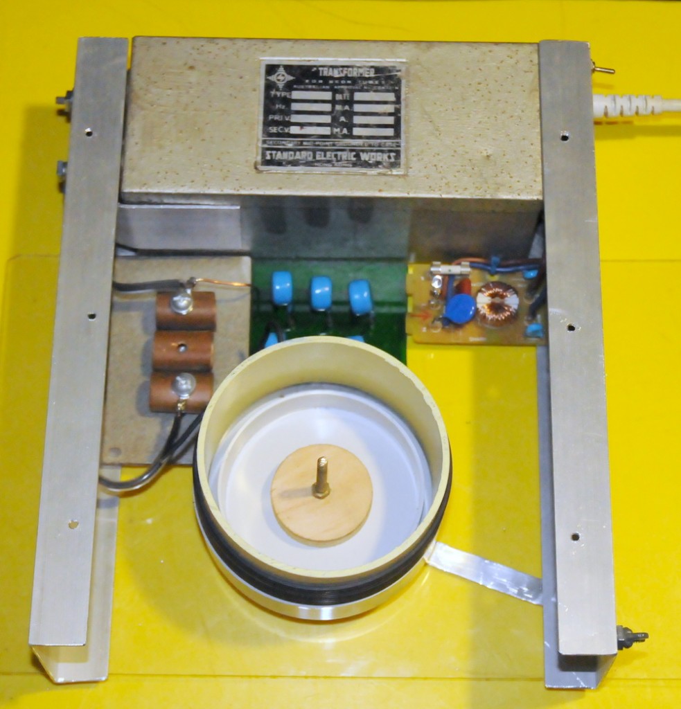 Briefcase Tesla coil internals with neon sign transformer at rear, copper pipe static spark gap at left, blue capacitors centre and mains suppression circuit at rear. 